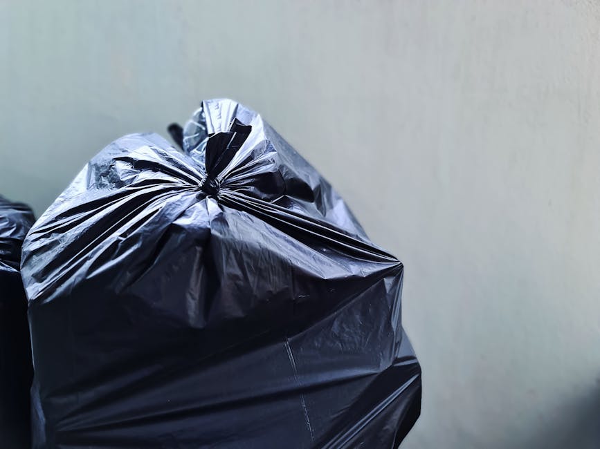 A sanitation worker dressed in a green uniform and safety helmet is standing on the driver’s side of a rear-loading rubbish collection truck parked on a residential street. The truck, which appears to